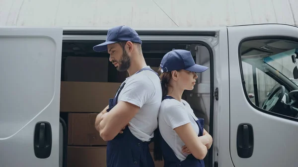 Vista lateral de los mensajeros de pie espalda con espalda cerca del coche con cajas de cartón al aire libre - foto de stock