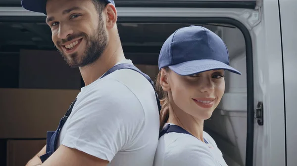 Mensajeros sonrientes mirando a la cámara mientras están de pie de espaldas cerca del coche al aire libre - foto de stock