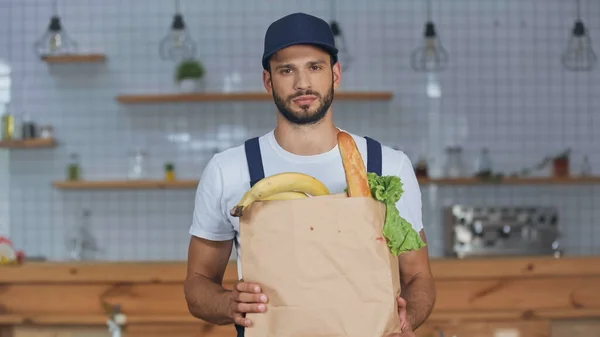 Zusteller blickt in Kamera, während er Paket mit Lebensmitteln zu Hause hält — Stockfoto