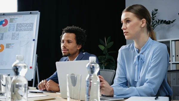 Jóvenes colegas de negocios interracial sentados cerca del ordenador portátil y botellas de agua en la sala de reuniones - foto de stock