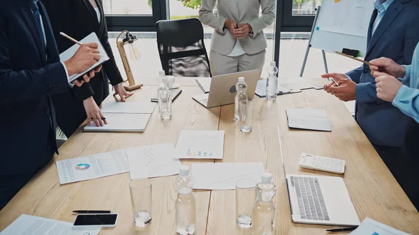 Partial view of multiethnic business people near desk with gadgets and documents in meeting room — Stock Photo