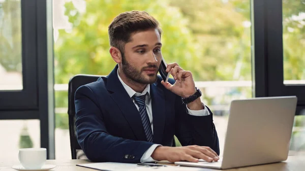 Giovane uomo d'affari che guarda il portatile durante la conversazione sul telefono cellulare — Foto stock