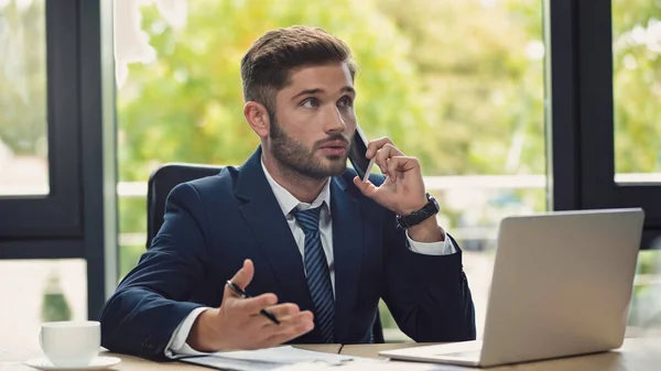 Thoughtful businessman talking on mobile phone near laptop and coffee cup — Stock Photo