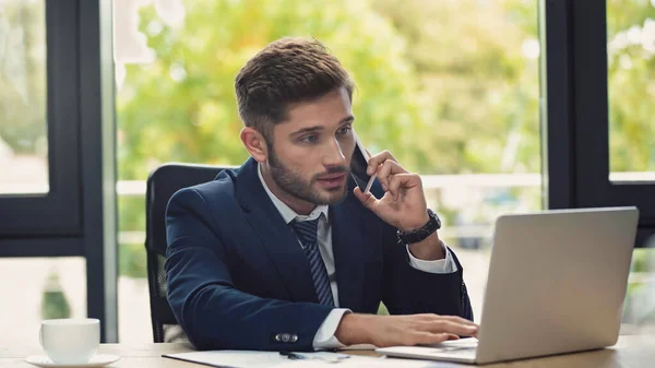 Joven hombre de negocios barbudo utilizando el ordenador portátil mientras llama al teléfono celular en el lugar de trabajo - foto de stock