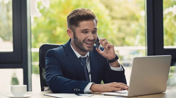 Alegre hombre de negocios hablando en el teléfono móvil cerca de portátil y taza de café - foto de stock