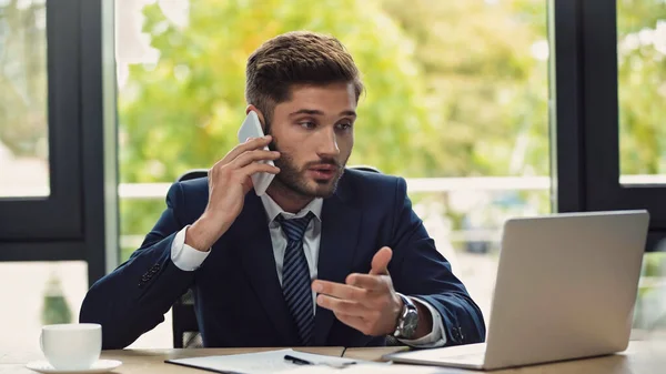 Joven empresario mirando a la computadora portátil mientras habla en el teléfono inteligente en la oficina - foto de stock