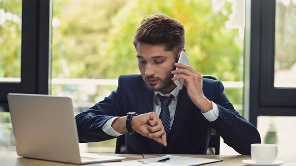 Geschäftsmann schaut auf Armbanduhr, während er in Laptopnähe mit dem Smartphone spricht — Stockfoto