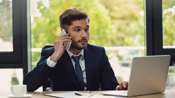 Jungunternehmer spricht auf Smartphone in der Nähe von Laptop und Kaffeetasse — Stockfoto