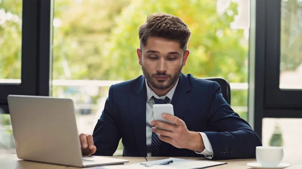 Jungunternehmer schaut auf Handy, während er im Büro am Laptop arbeitet — Stockfoto