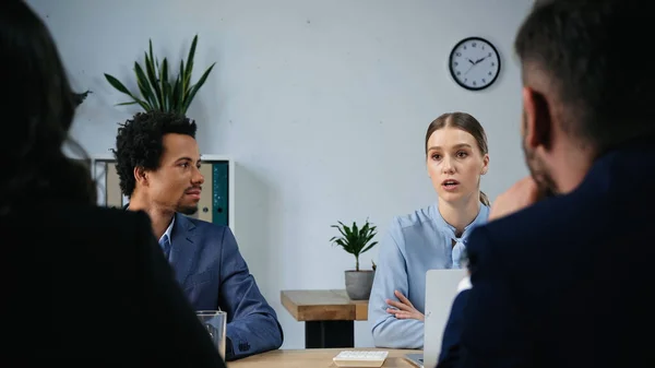 Joven mujer hablando cerca africano americano colega y borrosa negocios socios - foto de stock