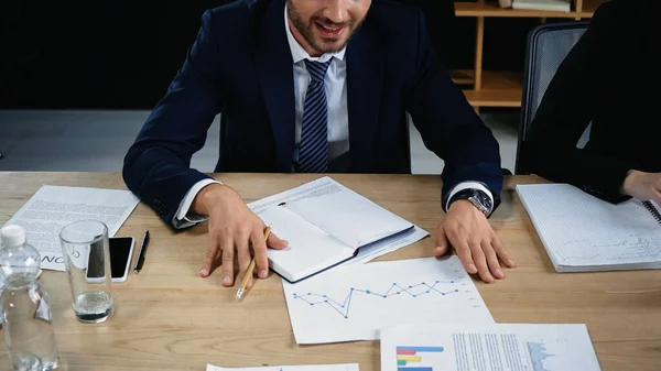 Cropped view of businessman in formal wear near empty notebook and graphs on desk — Stock Photo