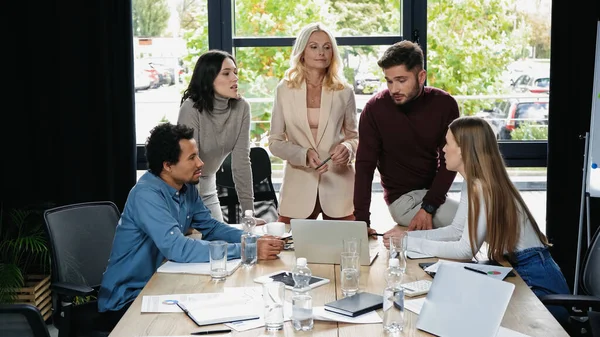 Colegas de negocios multiétnicos hablando cerca de escritorio con gadgets y documentos en la sala de reuniones - foto de stock