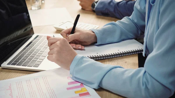 Cropped view of businesswoman writing in notebook near graphs and laptop with blank screen — Stock Photo