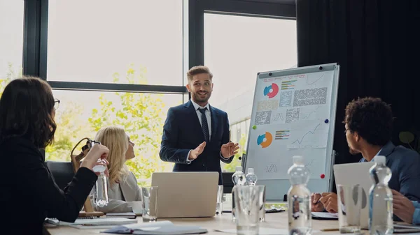 Smiling man talking near flipchart during business meeting with interracial colleagues — Stock Photo