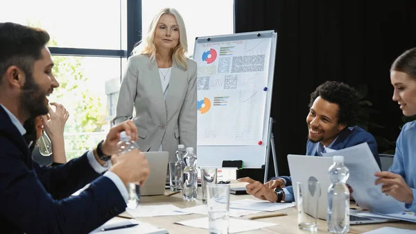 Middle aged businesswoman standing near smiling multiethnic managers in meeting room — Stock Photo