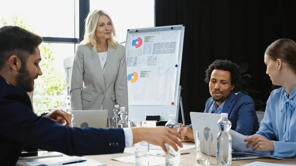 Smiling middle aged businesswoman standing near interracial colleagues talking on business meeting — Stock Photo