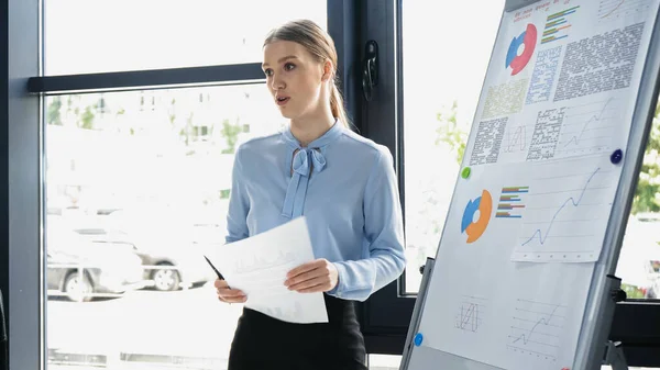 Young businesswoman holding document and talking near flip chart with graphs in office — Stock Photo