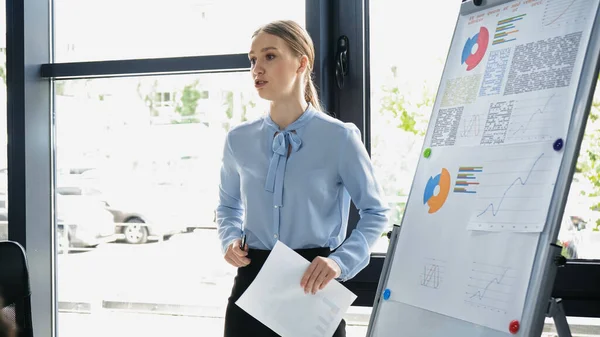 Giovane donna d'affari che parla vicino flip chart con infografica in ufficio — Foto stock