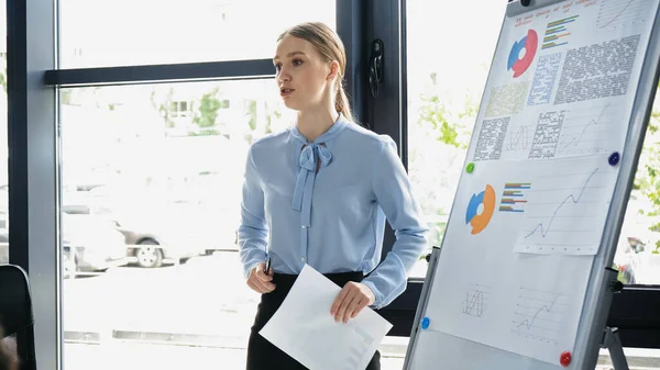 Young businesswoman talking near flip chart with infographics in office — Stock Photo