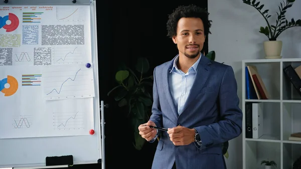 Joven hombre de negocios afroamericano sosteniendo pluma cerca de rotafolio con infografías - foto de stock