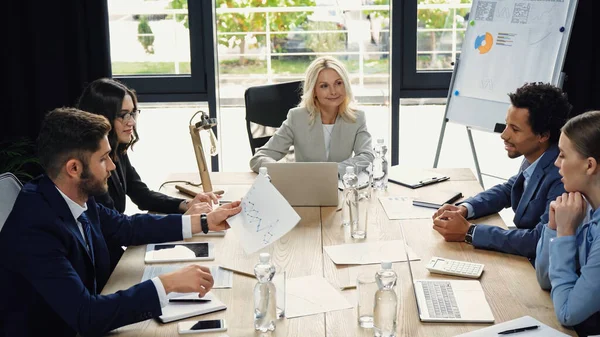 Young businessman holding infographics during meeting with multiethnic colleagues — Stock Photo