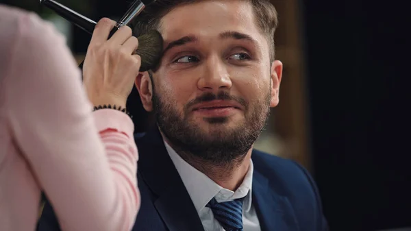 Maquillador borroso aplicando polvo facial al hombre sonriente en ropa formal - foto de stock