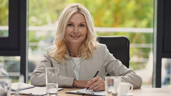 Femme d'affaires souriante tenant stylo tout en regardant la caméra près de verres d'eau flous — Photo de stock