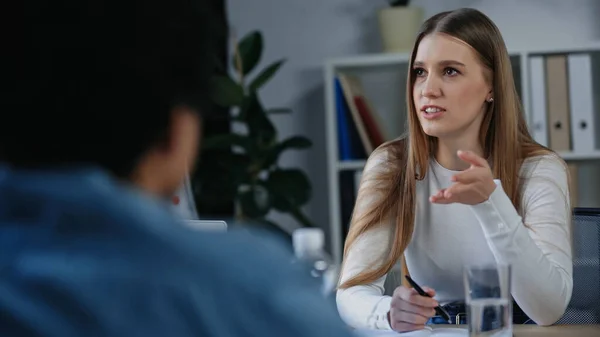 Joven empresaria señalando con la mano al borroso colega afroamericano - foto de stock