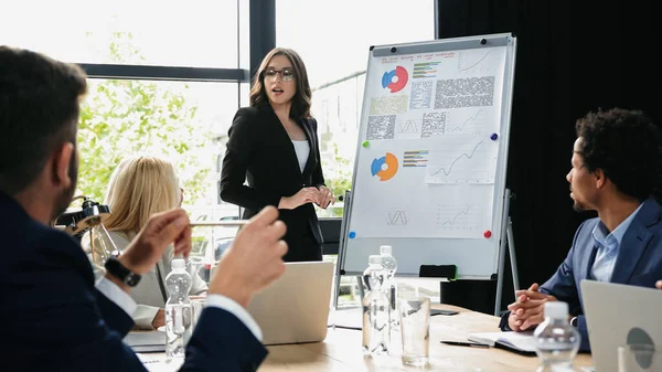 Young woman in eyeglasses talking near flipchart on business meeting with colleagues — Stock Photo
