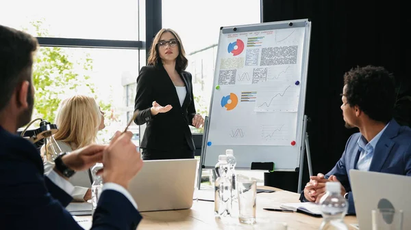 Empresária em óculos falando perto de flip chart com gráficos em reunião com colegas inter-raciais — Fotografia de Stock