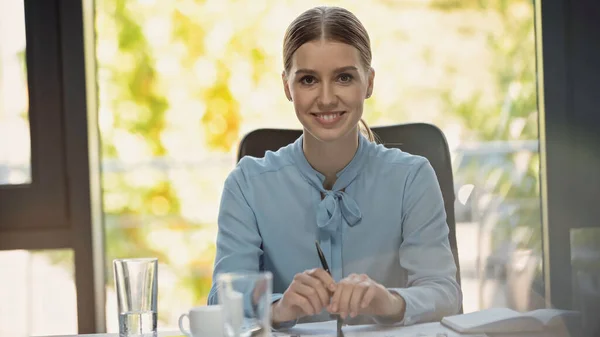 Cheerful businesswoman with pen looking at camera near blurred glasses in meeting room — Stock Photo