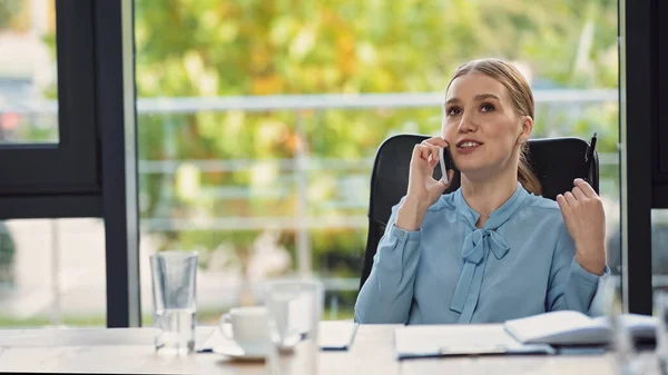 Sonriente mujer de negocios hablando en el teléfono celular cerca borrosa portátil y bebidas — Stock Photo