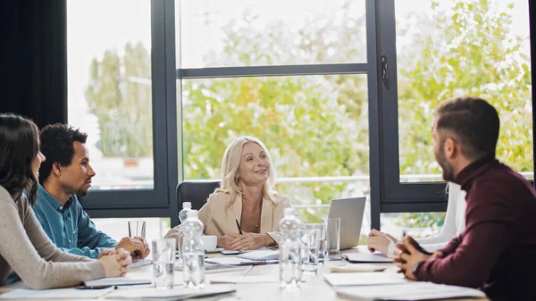Positive Geschäftsfrau mittleren Alters im Gespräch mit jungen multiethnischen Managern während eines Treffens — Stockfoto