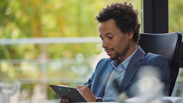 Joven empresario afroamericano escribiendo en portapapeles en primer plano borroso - foto de stock