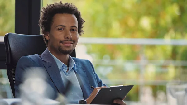 Happy african american businessman with clipboard and pen looking at camera in office — Stock Photo