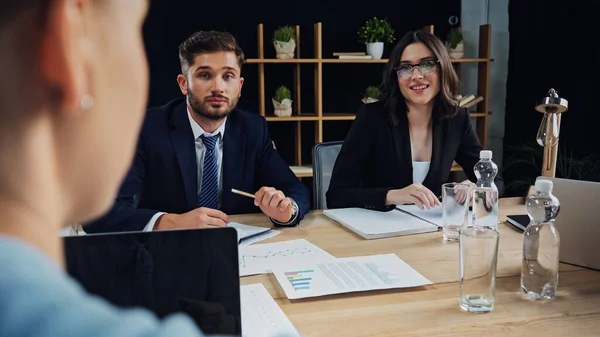 Young business people looking at blurred colleague near documents and laptop with blank screen — Stock Photo