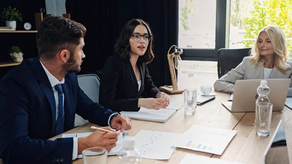 Joven mujer de negocios en anteojos hablando con colegas en la sala de reuniones - foto de stock