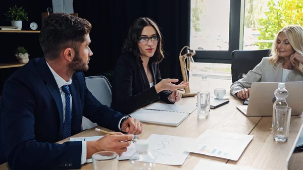 Femme d'affaires dans les lunettes pointant avec la main tout en parlant à des collègues lors de la réunion — Photo de stock