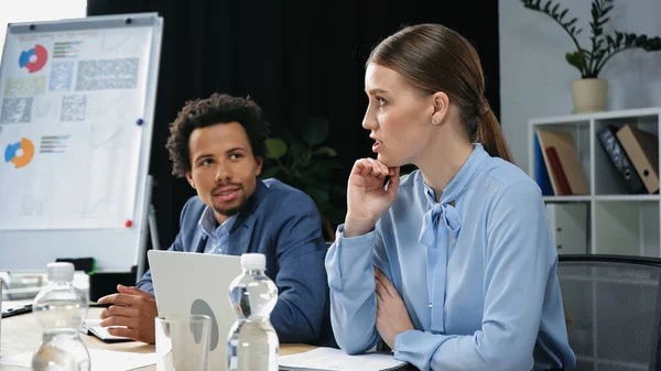 Pensativa mujer de negocios hablando cerca de un colega afroamericano y portátil en la sala de reuniones - foto de stock