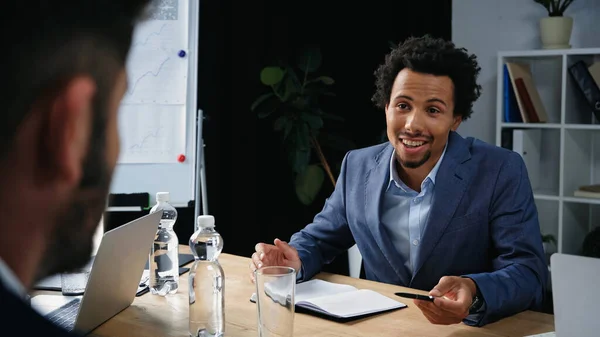 Sonriente hombre de negocios afroamericano apuntando con bolígrafo mientras habla con un colega borroso - foto de stock