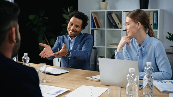 Happy african american businessman pointing at blurred colleague during meeting — Stock Photo