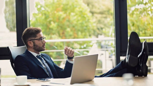 Pensive businessman sitting near laptop with legs on desk — Stock Photo