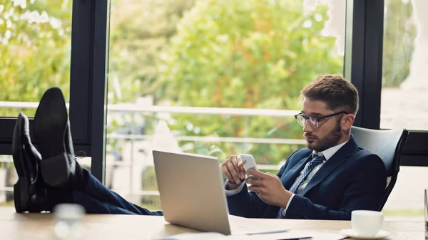 Hombre de negocios en gafas de vista sentado cerca de la computadora portátil con las piernas en el escritorio y escribiendo en el teléfono inteligente - foto de stock