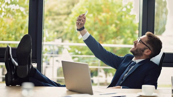 Alegre hombre de negocios tomando selfie mientras está sentado con piernas en el escritorio en el lugar de trabajo - foto de stock