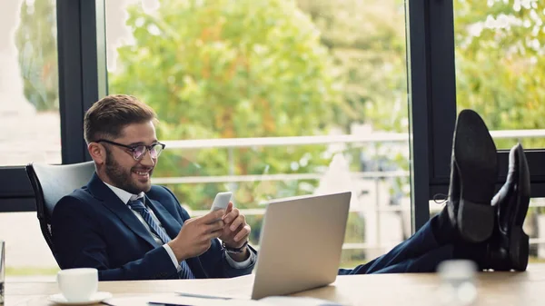 Uomo d'affari sorridente messaggistica sul telefono cellulare mentre seduto vicino al computer portatile con le gambe sulla scrivania — Foto stock