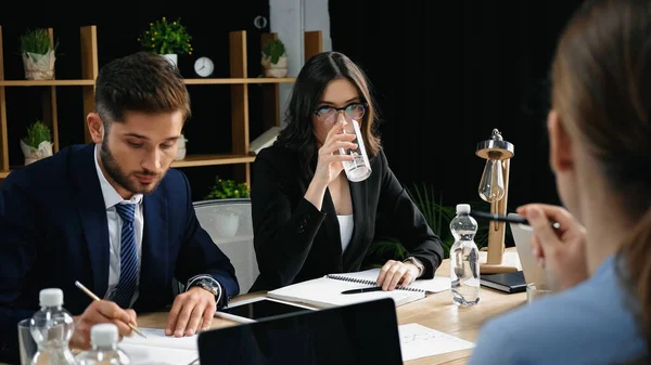 Mujer joven en anteojos agua potable durante reunión de negocios con colegas - foto de stock