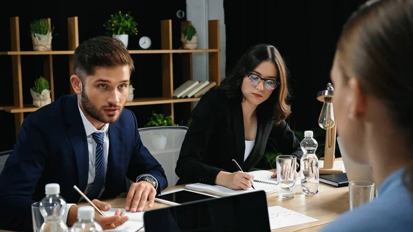 Young man talking to blurred colleague near businesswoman writing in notebook — Stock Photo