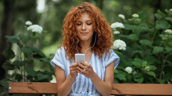 Cheerful redhead woman listening music in earphones and using smartphone in park — Stock Photo