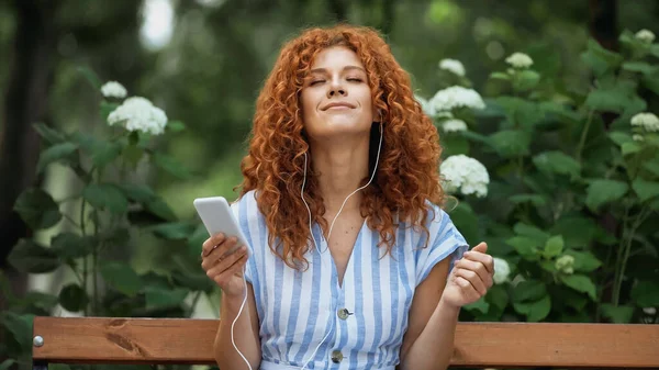 Довольная рыжая женщина с закрытыми глазами слушает музыку в наушниках и использует смартфон в парке — стоковое фото
