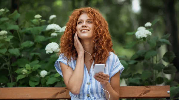 Positive redhead woman listening music in earphones and using smartphone in green park — Stock Photo